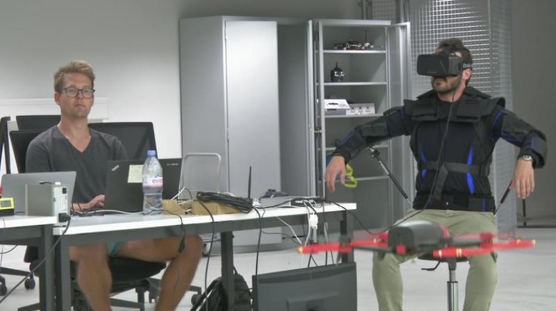 Robotics expert on the Ecole Polytechnique Federal de Lausanne, EPFL team, Matteo Macchini, right, displays the fly controls in a virtual reality scenario as he controls the flight of a drone in foreground, while observed by a fellow member of the team, July 23, 2018, in Lausanne, Switzerland. Scientists, engineers and robotics experts at the Swiss university have teamed up to develop a jacket that allows hands-free, torso-controlled flying of drones. (Ecole Polytechnique Federale De Lausanne via AP) MANDATORY DREDIT