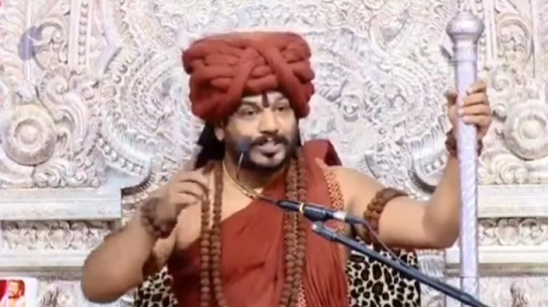 Nithyananda, who is sporting a bear, rust-coloured robes and rudraksh around his neck and hands as well as holding a silver staff, is heard making tall claims and promises before audience that is cheering and applauding wildly after his statements. (Youtube Screengrab/ DR K Tv)