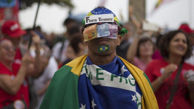 A man poses for the photo with his face covered with money and by a sign that reads in Portuguese Temer Out. (Photo: AP)